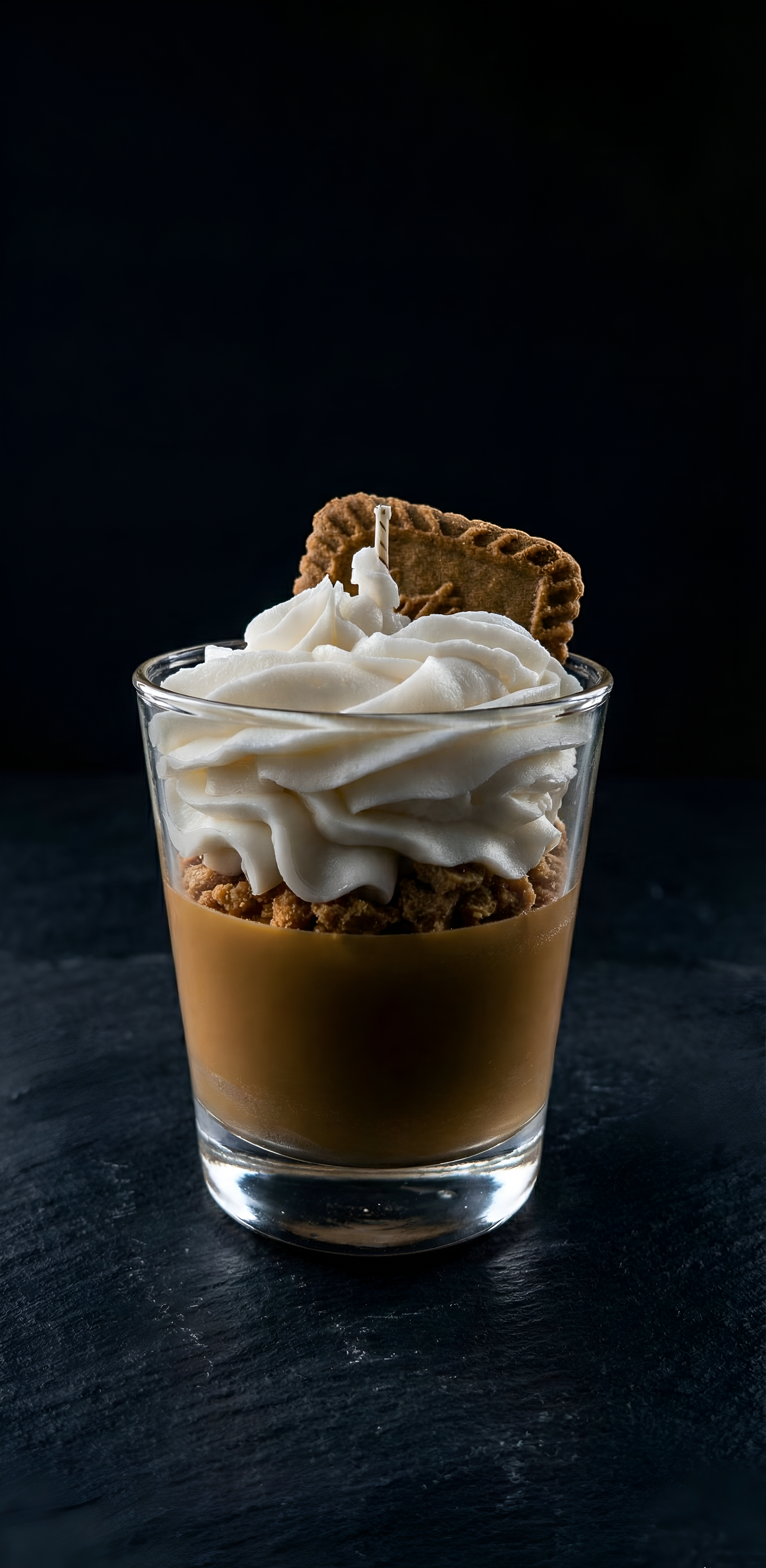 Gros plan sur une bougie dessert avec des couches de cire caramel et chantilly surmontée d'un biscuit, présentée sur une surface en ardoise noire sombre pour un contraste élégant