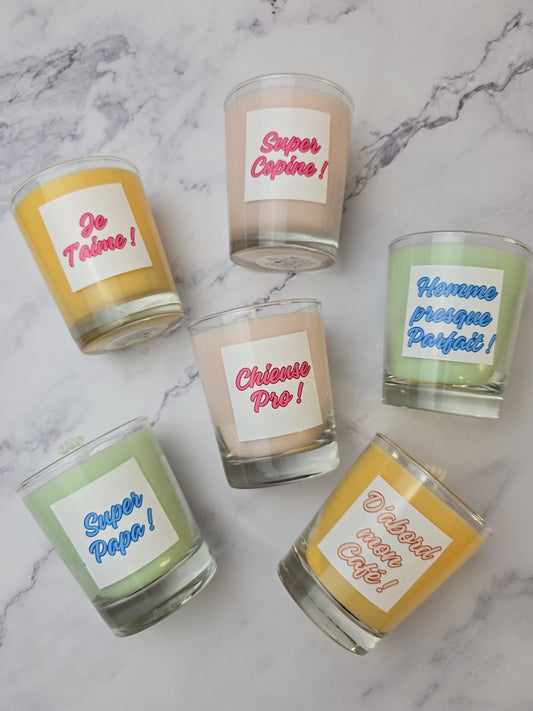 Bougies personnalisées à message amusant en verrines colorées sur fond marbré blanc. Les étiquettes montrent des citations françaises : "Je T'aime", "Super Copine", "Homme presque Parfait !", "Super Papa !", "Chieuse Pro !", "D'abord mon Café !".