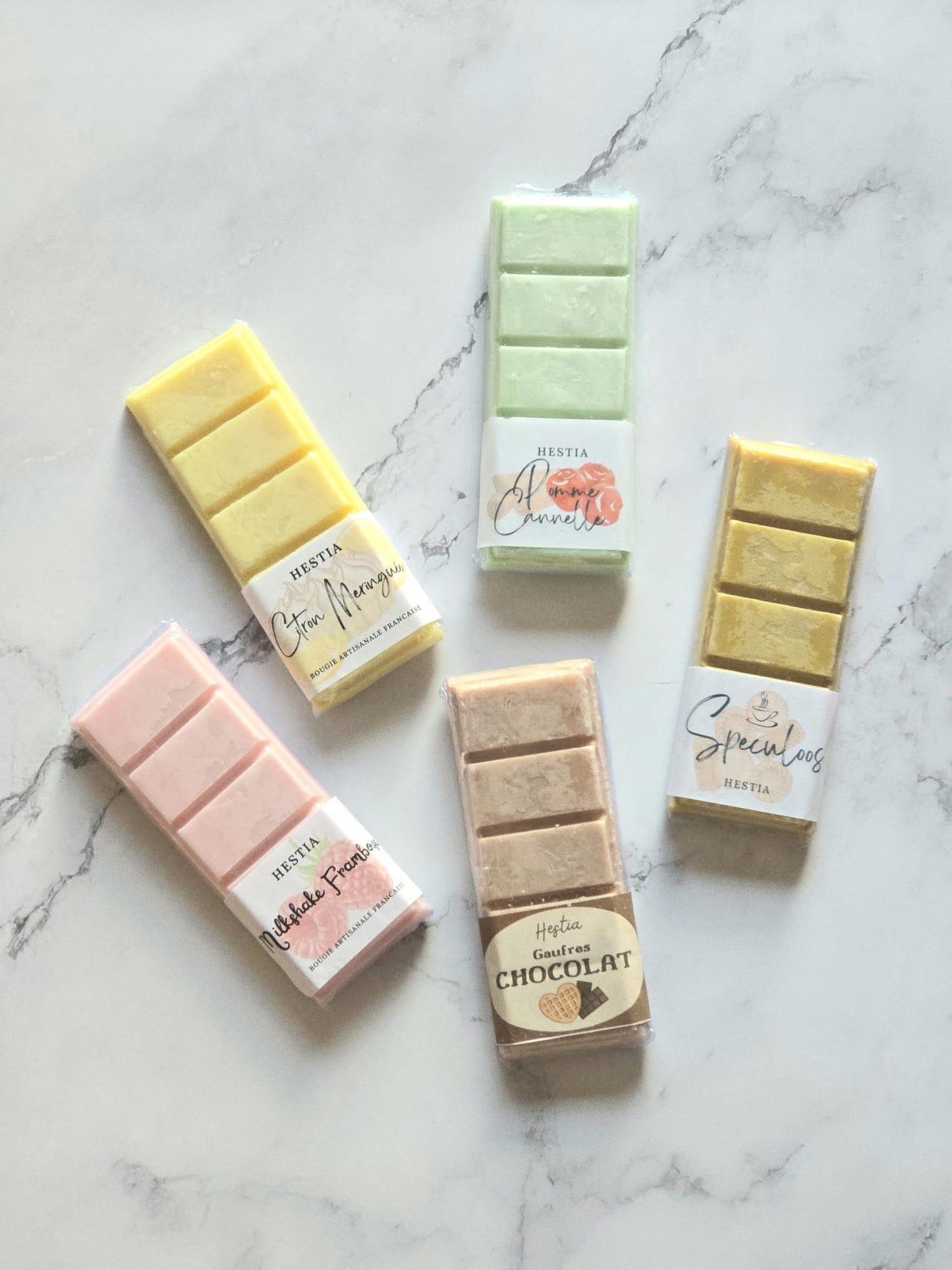 Quatre tablettes de fondants parfumés artisanaux en cire de soja de différentes couleurs (jaune, rose, vert, marron) avec étiquettes de parfums gourmands. Vue d'ensemble des carrés de cire sur un fond de marbre blanc.