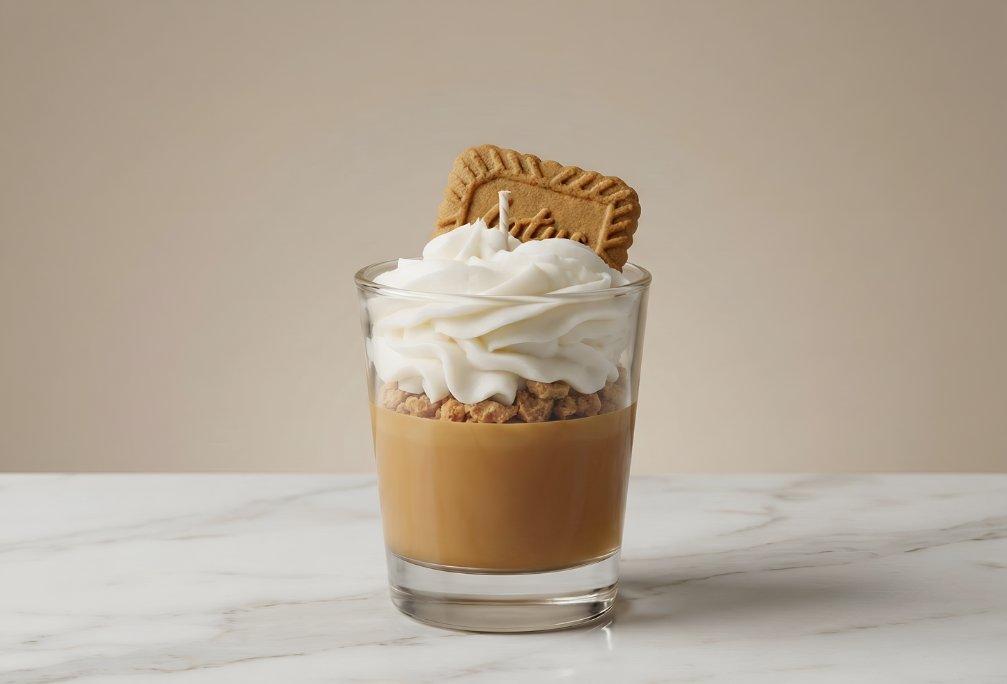 Une bougie gourmande imitant un dessert en couches avec un biscuit Lotus sur le dessus, posée sur une surface en marbre blanc avec un fond beige neutre, style photographie de produit épurée