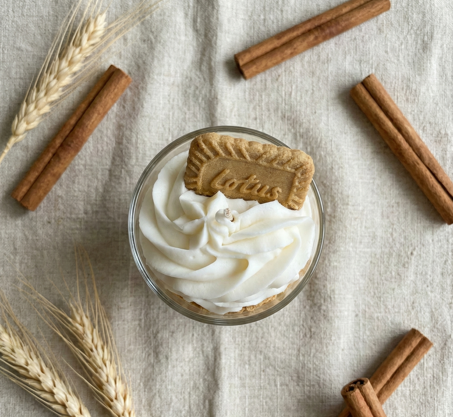 Vue de dessus d'une bougie artisanale blanche ornée d'un biscuit, disposée au centre d'un tissu en lin et entourée esthétiquement de bâtons de cannelle et d'épis de blé