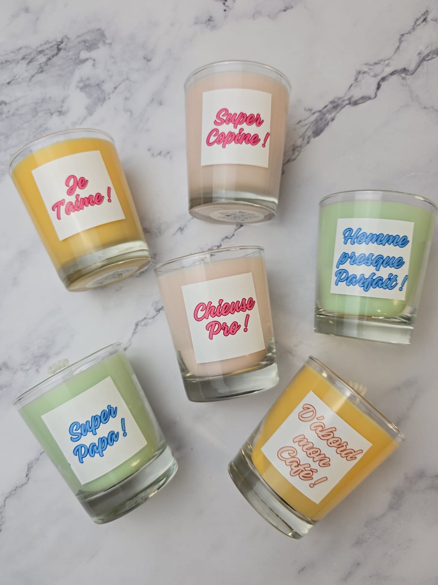 Bougies personnalisées à message amusant en verrines colorées sur fond marbré blanc. Les étiquettes montrent des citations françaises : "Je T'aime", "Super Copine", "Homme presque Parfait !", "Super Papa !", "Chieuse Pro !", "D'abord mon Café !".
