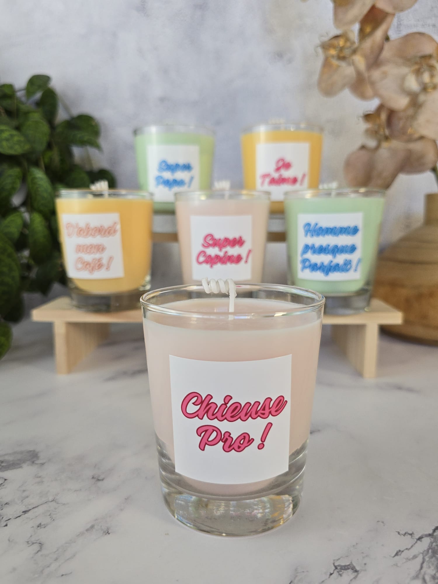 Bougies personnalisées à message amusant en verrines colorées sur fond marbré blanc. Les étiquettes montrent des citations françaises : "Je T'aime", "Super Copine", "Homme presque Parfait !", "Super Papa !", "Chieuse Pro !", "D'abord mon Café !".